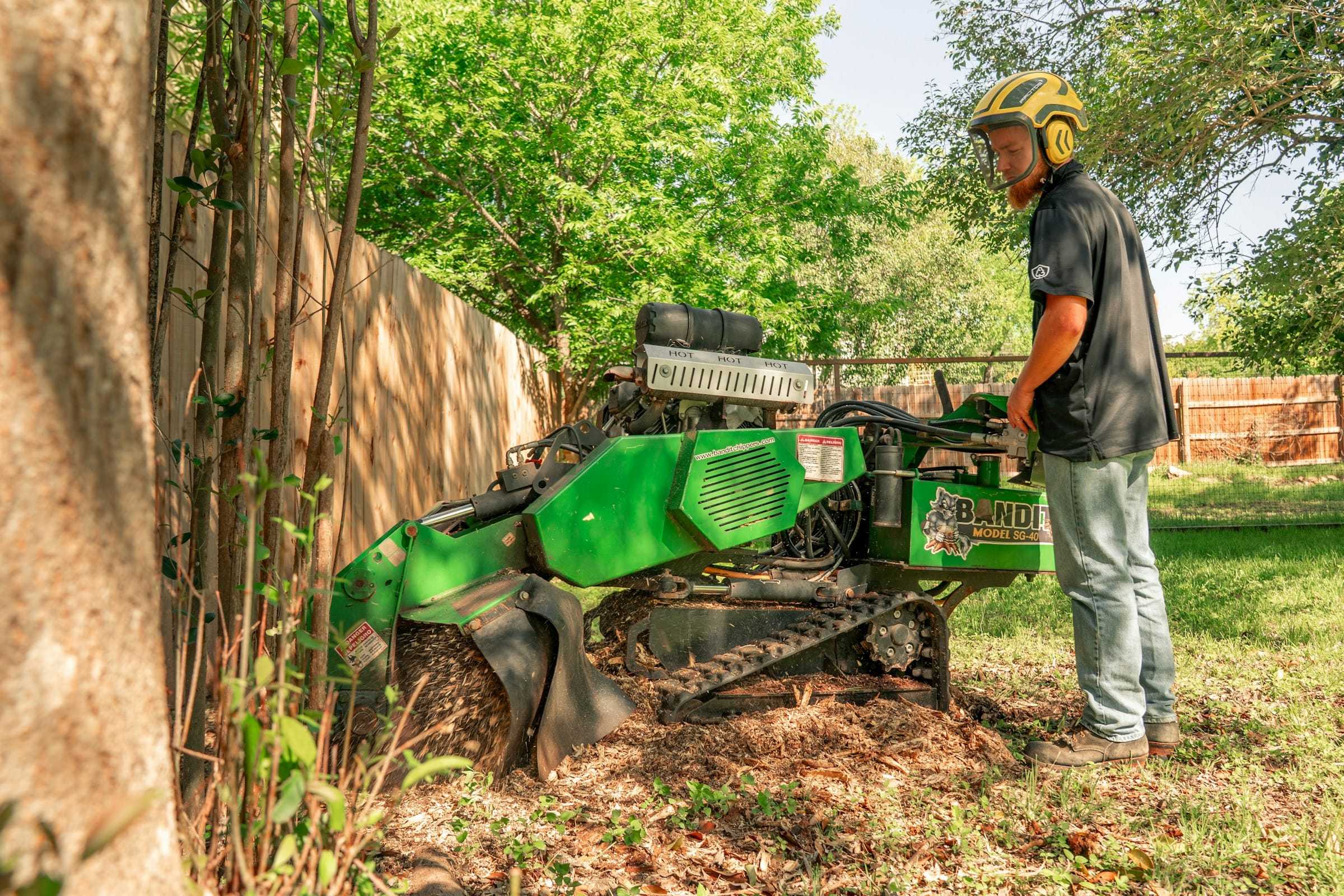 Operator in a helmet running a compact stump grinder in a fenced backyard